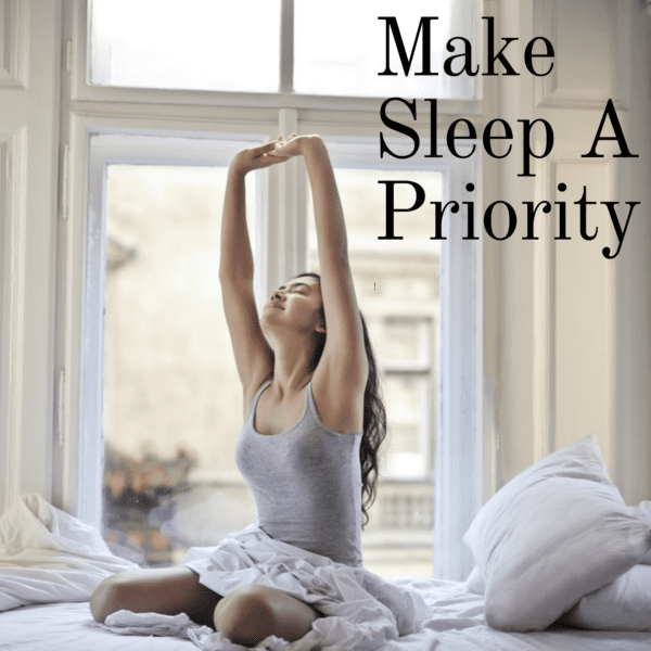 A woman sitting up in bed stretching overhead and smiling and looking refreshed after a good night's sleep. Make sleep a priority, quote.